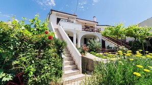 A two-story house with a staircase leading to a balcony, surrounded by lush greenery and flowering plants. - Apartments Puntica (Punat)