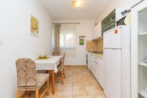 A compact kitchen with a dining area, featuring a table with chairs, a refrigerator, and a window with curtains. - Apartments Puntica (Punat)
