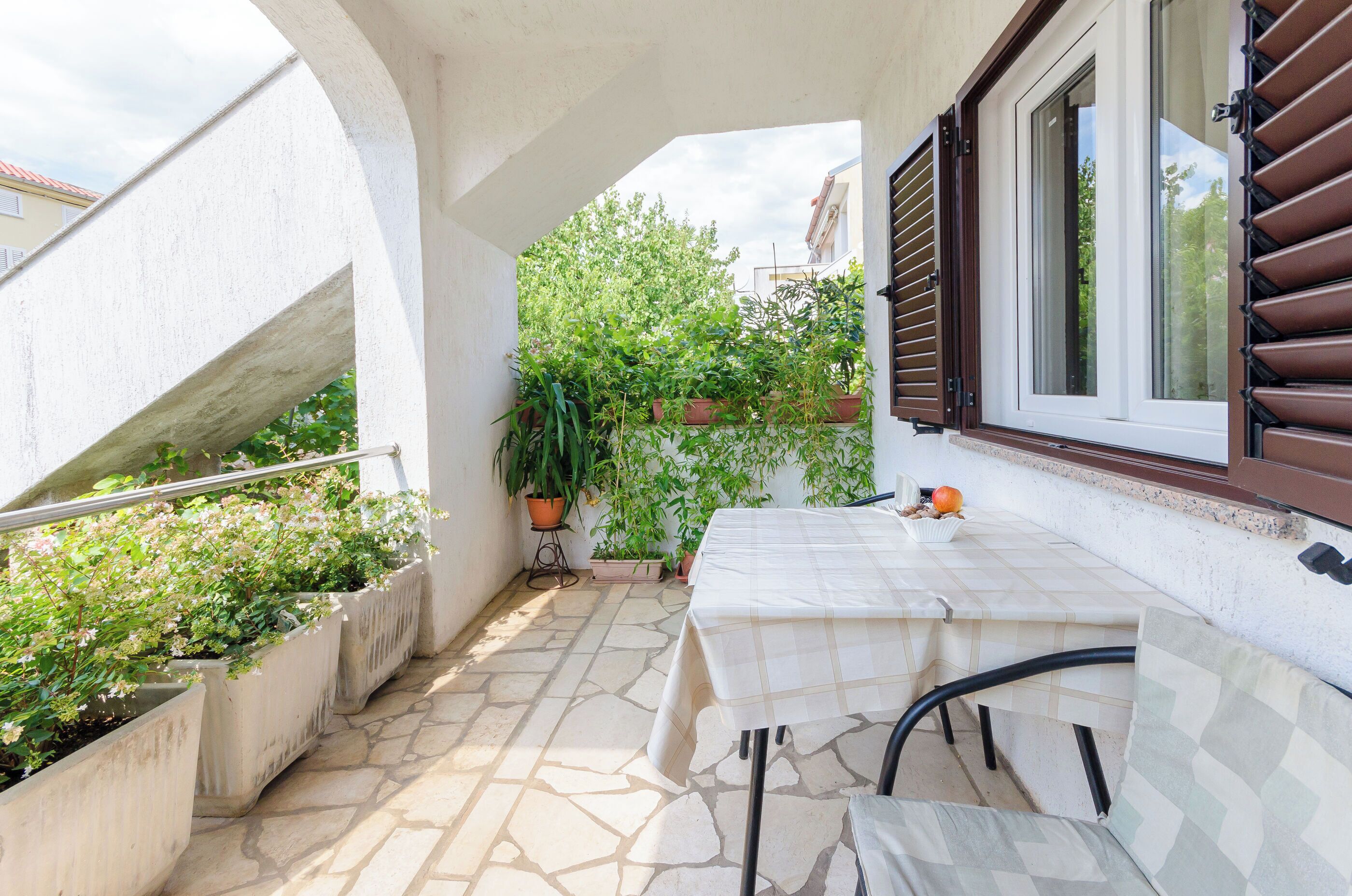 A balcony with a table, chairs, potted plants, and a window with shutters.