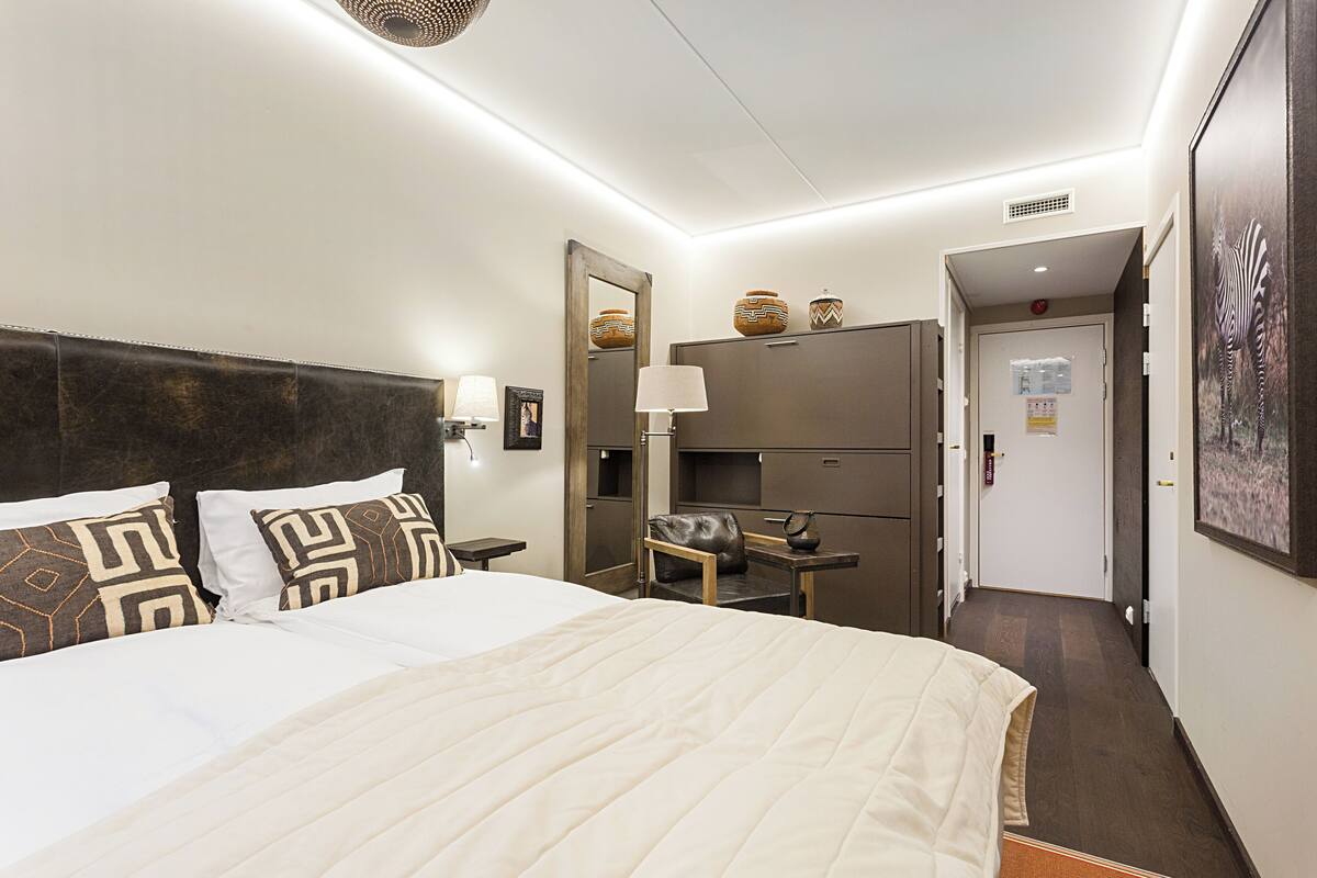 Quarto de hotel com uma cama grande, uma escrivaninha, uma cadeira, um espelho e uma porta que dá para outro quarto.