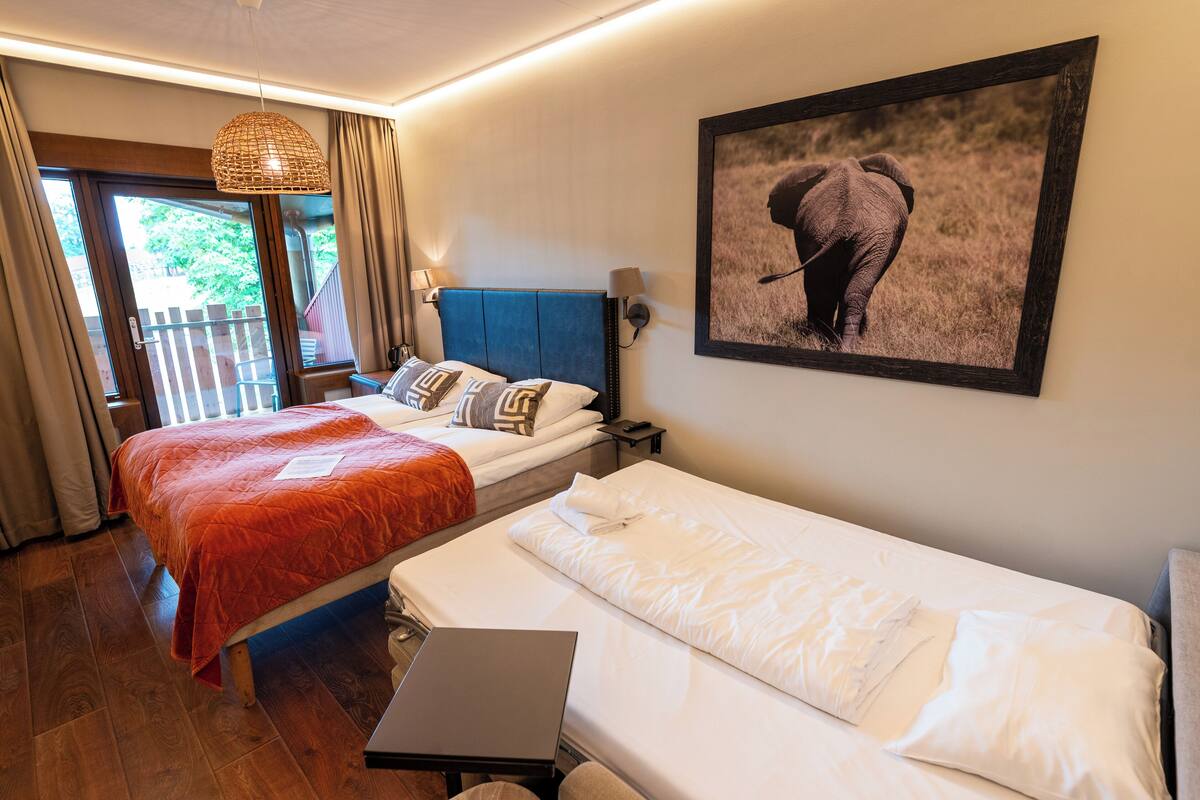 Quarto de hotel com cama, escrivaninha, laptop, fotografia de um elefante e varanda com cortinas.