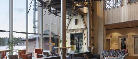 A spacious indoor area with a high ceiling, large windows, and a view of the outdoors. There are multiple seating areas with sofas and chairs, and a staircase with a rope bridge.