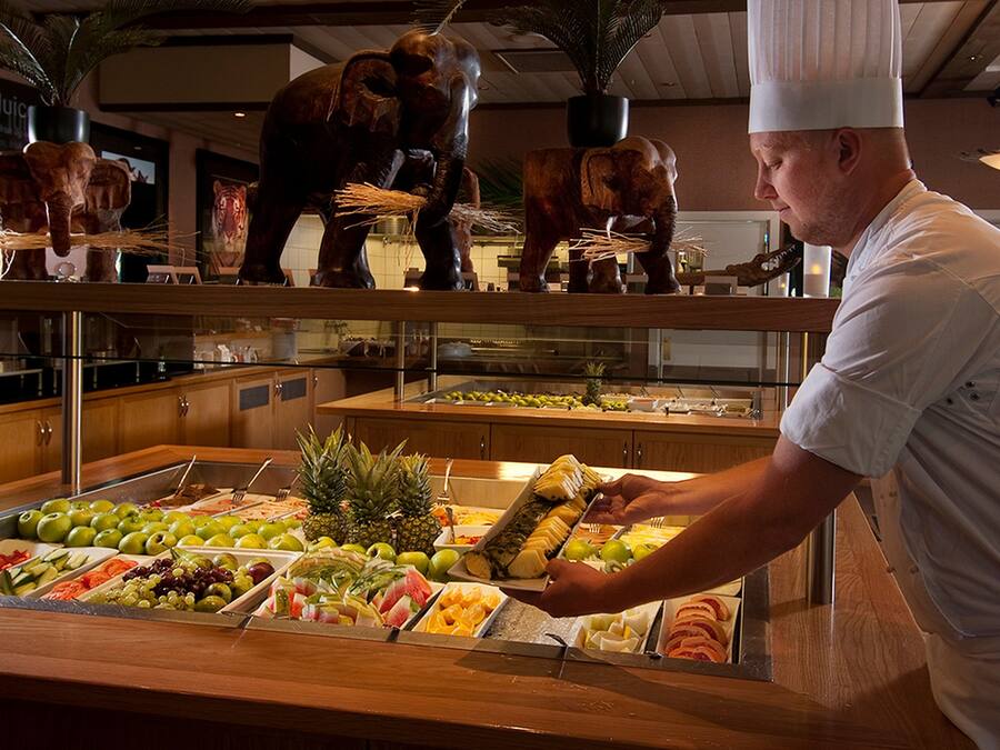 Un bufé con diversas frutas y verduras, un chef sirviendo comida y esculturas decorativas de elefantes.