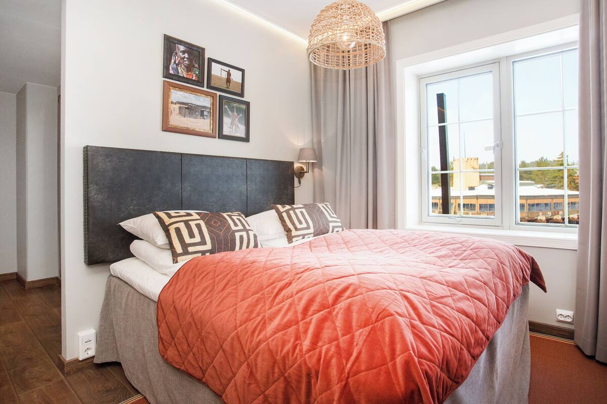 Cama bem arrumada com cabeceira estampada, luminária de cabeceira e janela com cortinas.