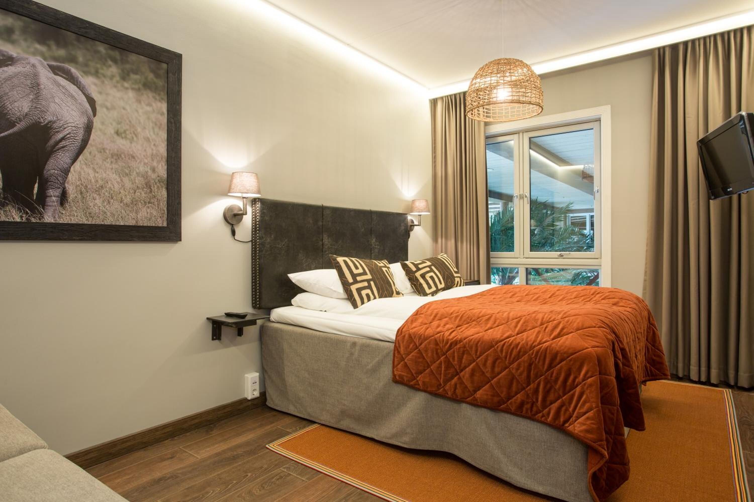 A hotel room with a bed, a nightstand, a wall-mounted TV, a window with curtains, and a picture of an elephant on the wall.