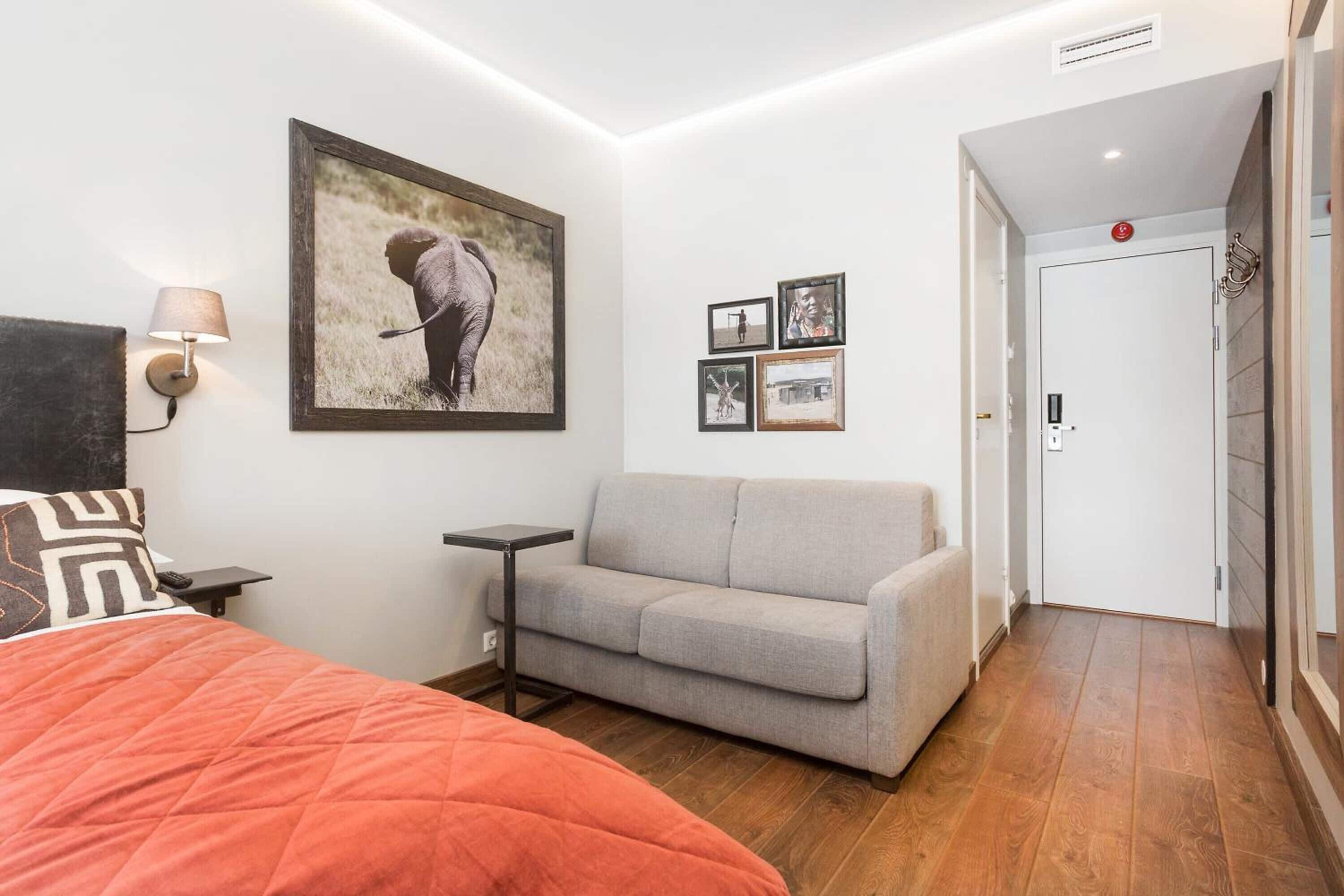 A hotel room with a bed, a grey sofa, a small table, a wall-mounted lamp, and a framed picture of an elephant.
