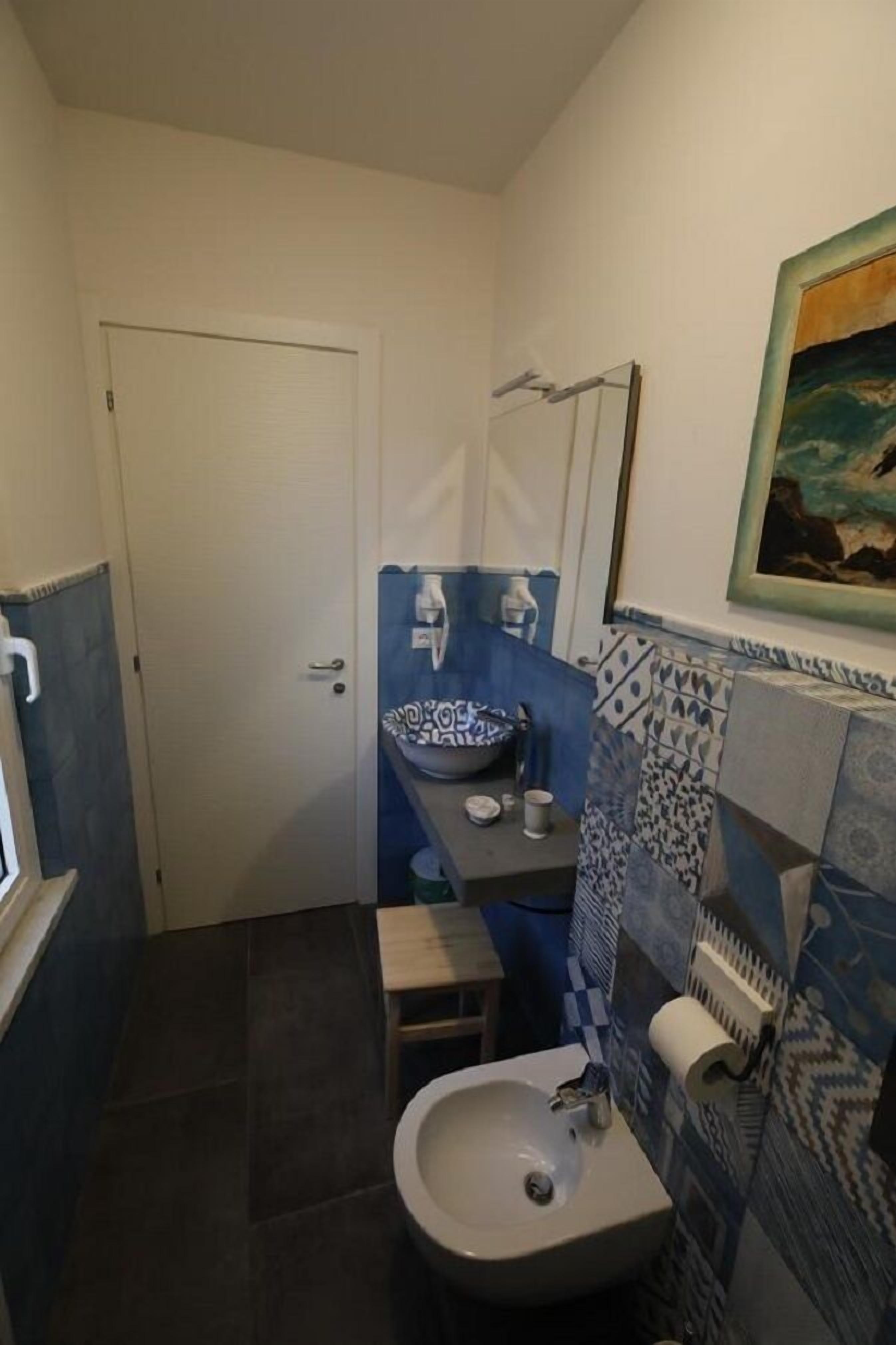 A compact bathroom with a white door, a sink with a blue patterned basin, a small stool, and a toilet.