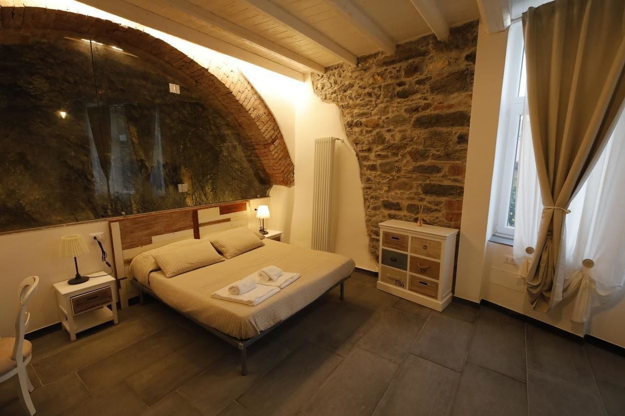 Un dormitori amb una finestra gran d'arc, un llit amb edredó blanc, una tauleta de nit amb una làmpada i un petit armari.