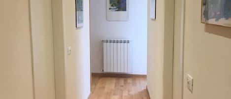 Un pasillo con suelo de madera, paredes blancas y un radiador. Hay cuadros enmarcados en las paredes y una puerta a la izquierda.