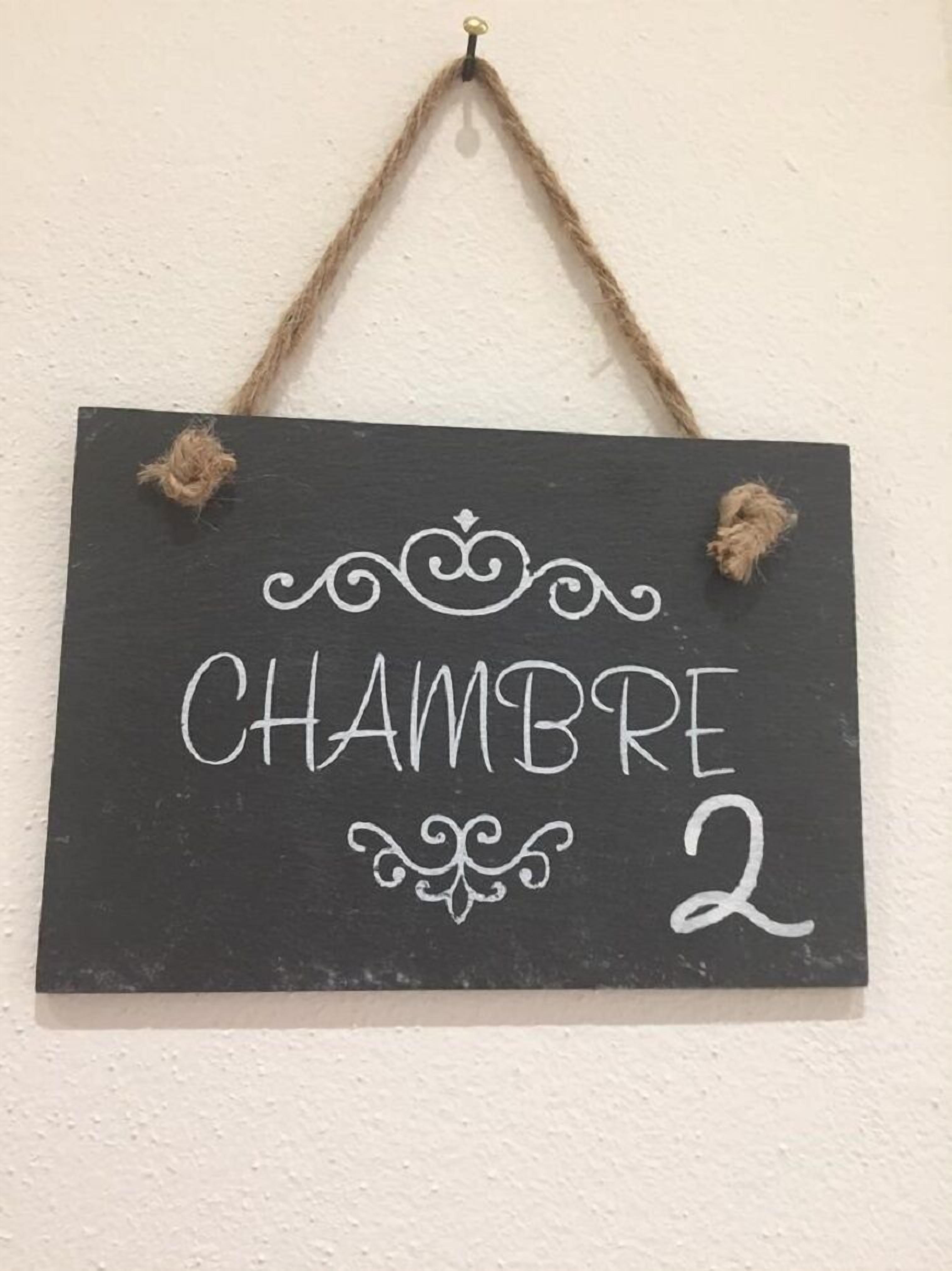 Un panneau noir avec le texte « CHAMBRE 2 » et des motifs décoratifs, accroché à un mur sur un fond simple et épuré.