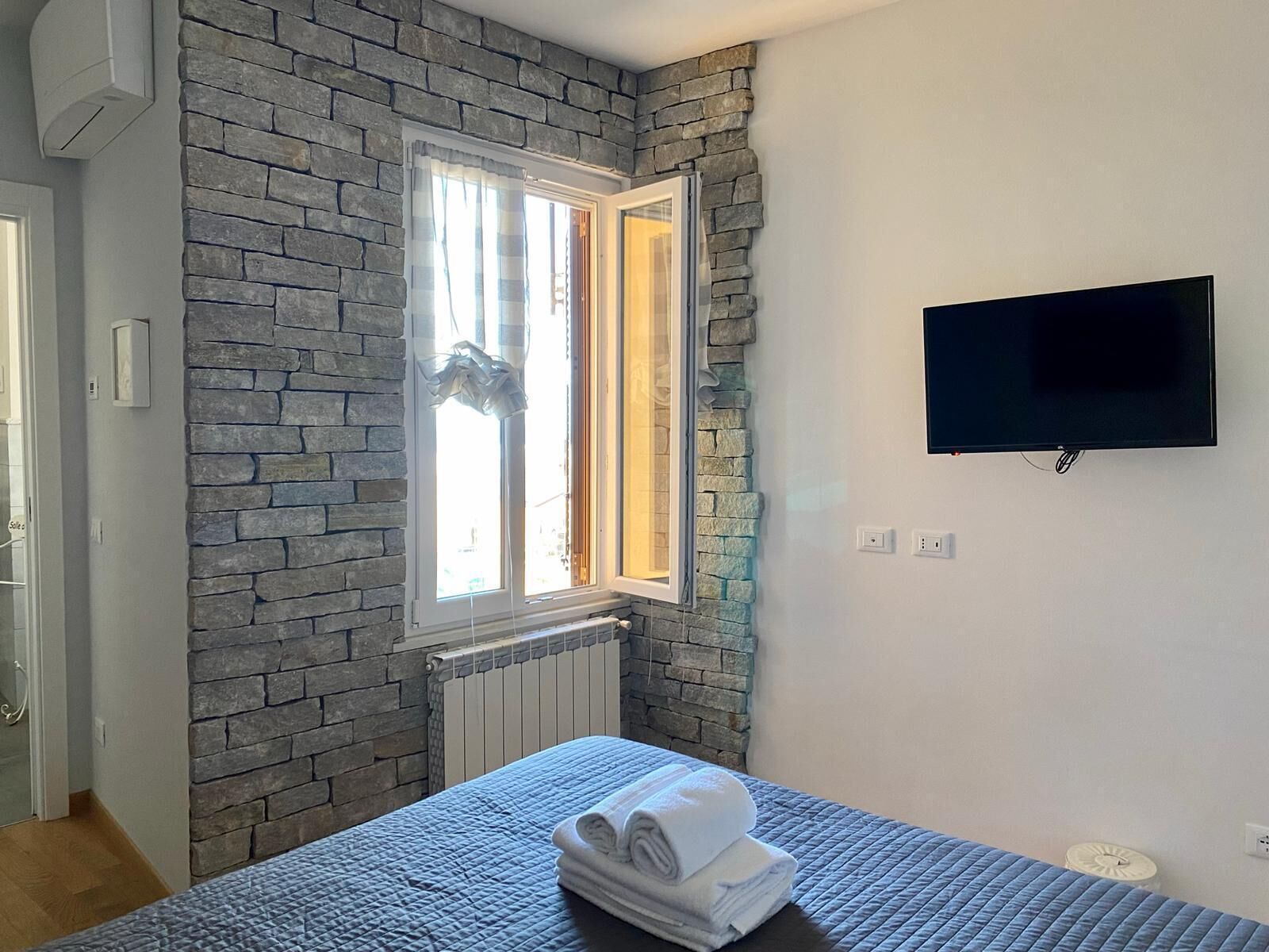 Une chambre avec un mur en pierre, une fenêtre avec des rideaux, un lit recouvert d’une couette bleue et un téléviseur fixé au mur.