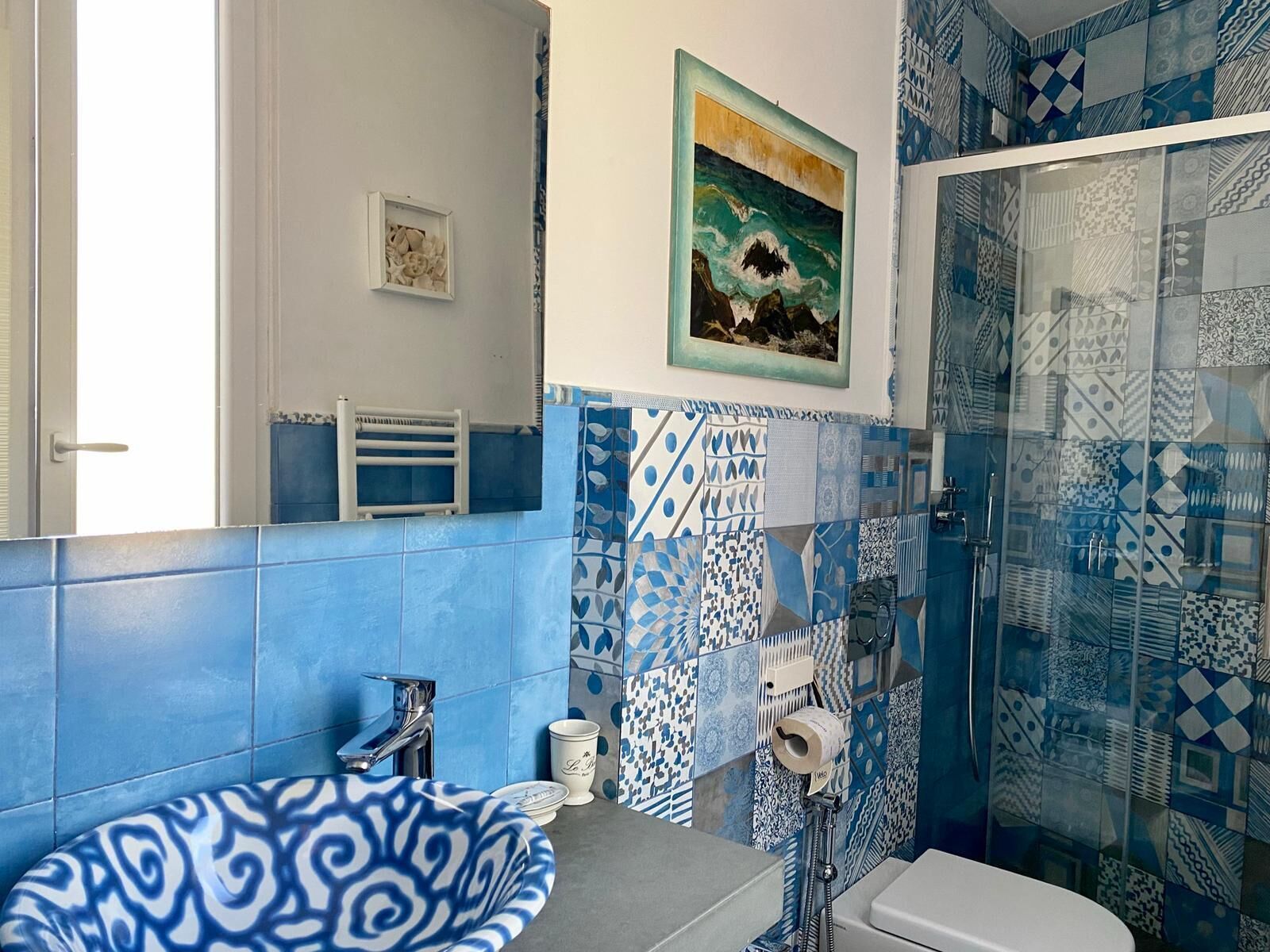 A bathroom with blue and white patterned tiles, a sink, a toilet, a shower with glass doors, and a framed painting on the wall.