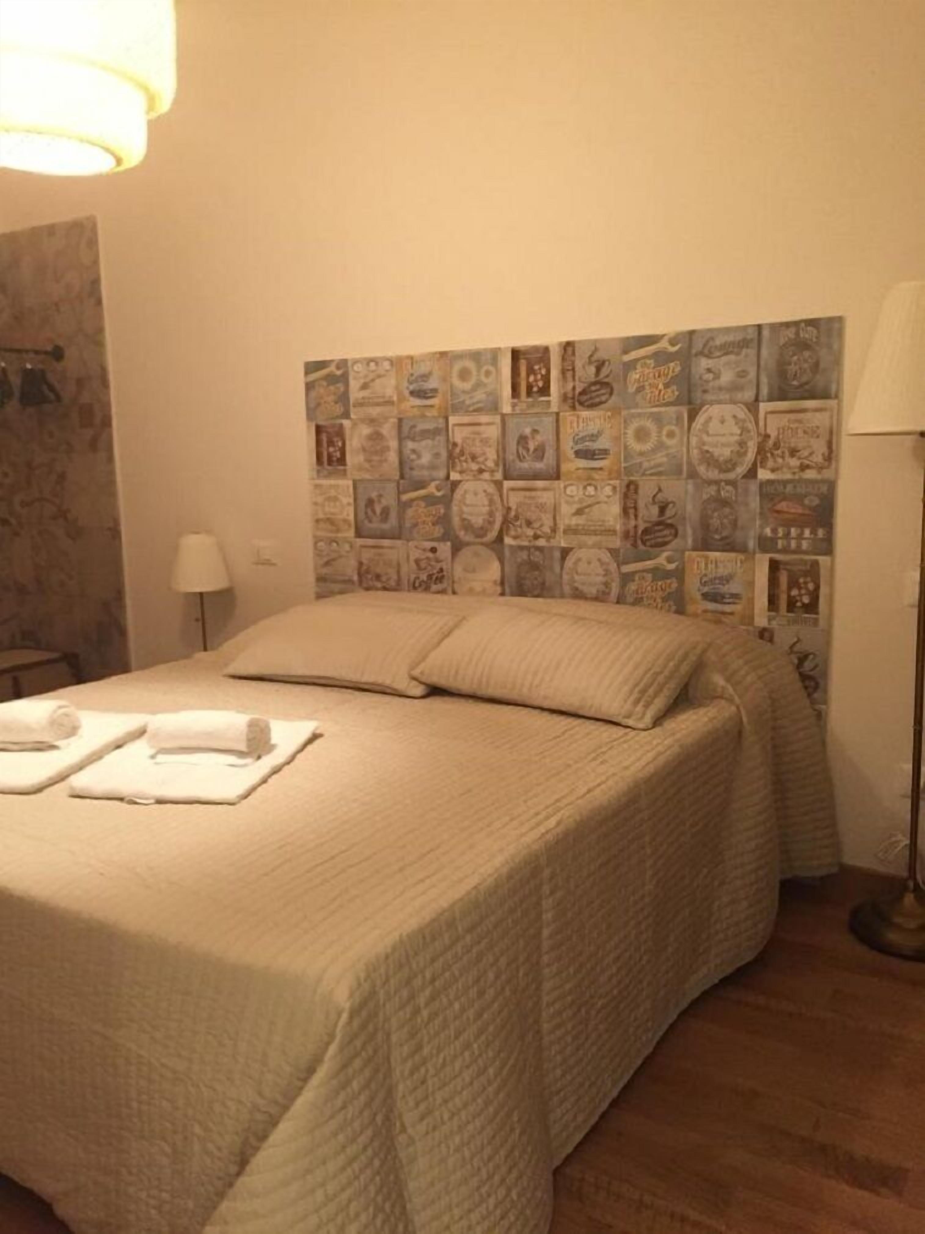 Quarto de hotel com uma cama, duas almofadas, uma mesinha de cabeceira, um abajur e uma parede decorada com cartazes antigos.