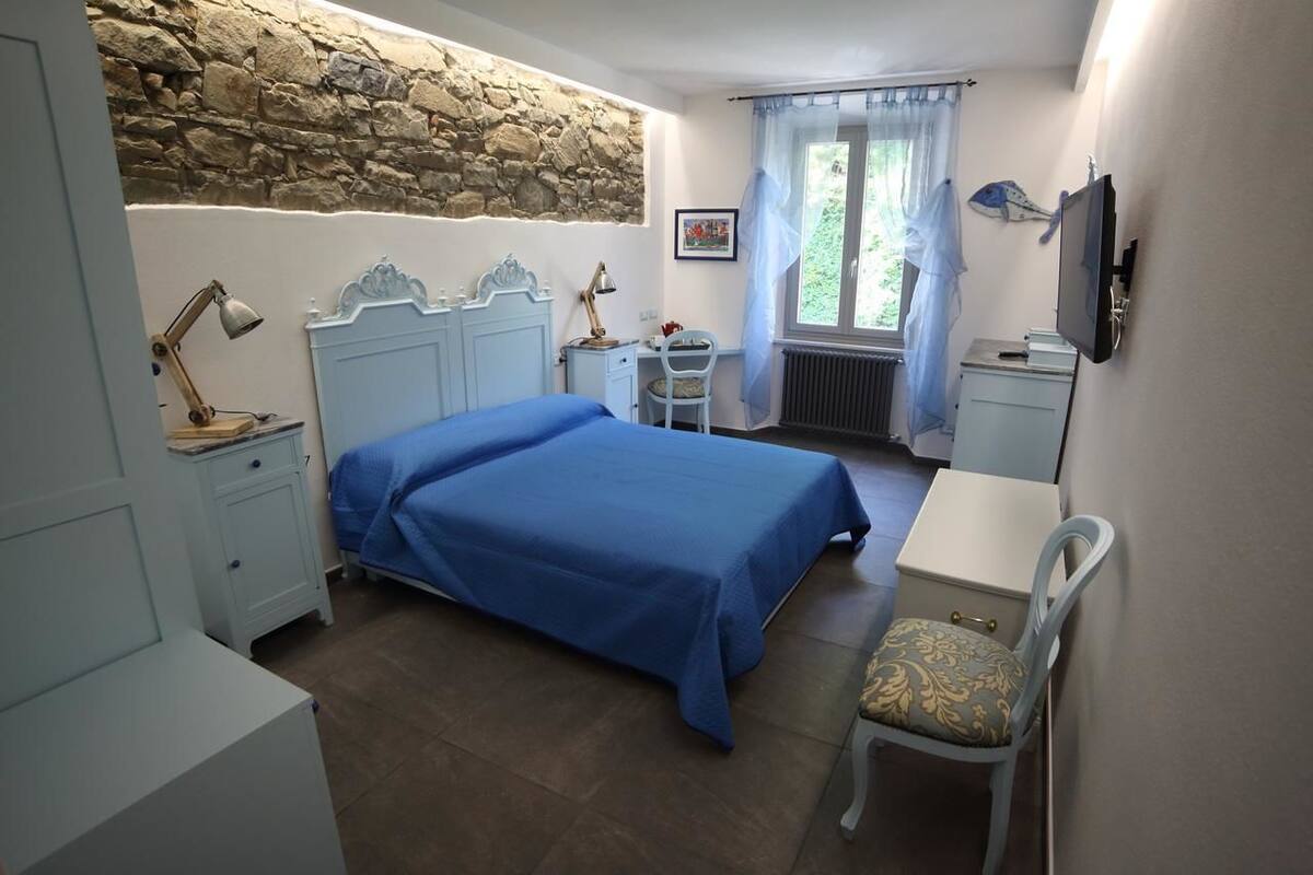 A bedroom with a stone wall, a bed with a blue bedspread, a desk with a chair, a radiator, and a television.