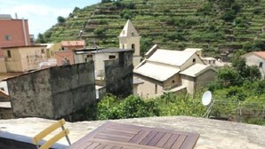 Una mesa de madera y dos sillas en una terraza en la azotea, con vistas a campos escalonados y edificios al fondo.