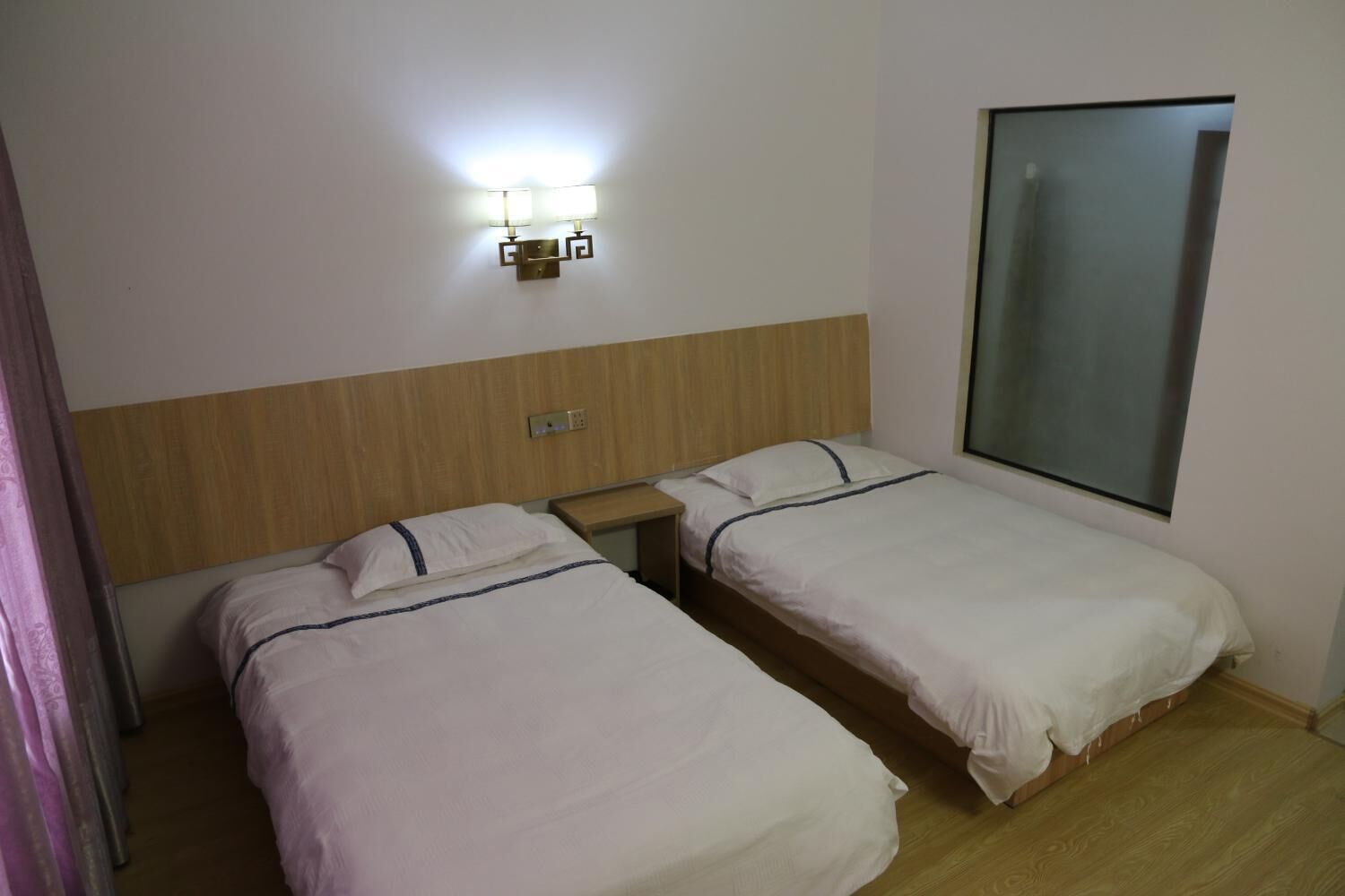 A hotel room with two single beds, a wooden headboard, a nightstand, and a wall-mounted light fixture.