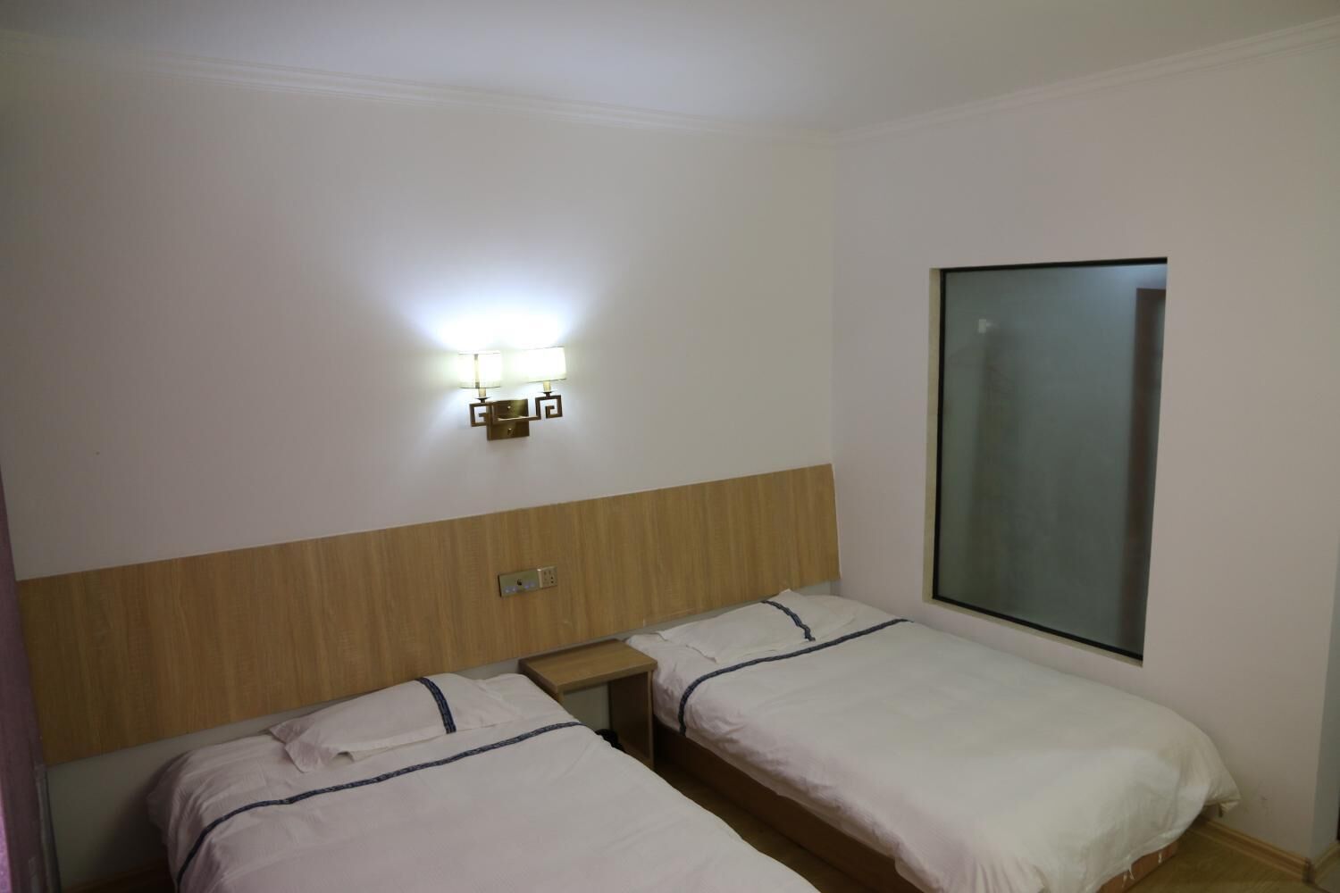 A hotel room with two single beds, a nightstand, a wall-mounted light fixture, and a large window.