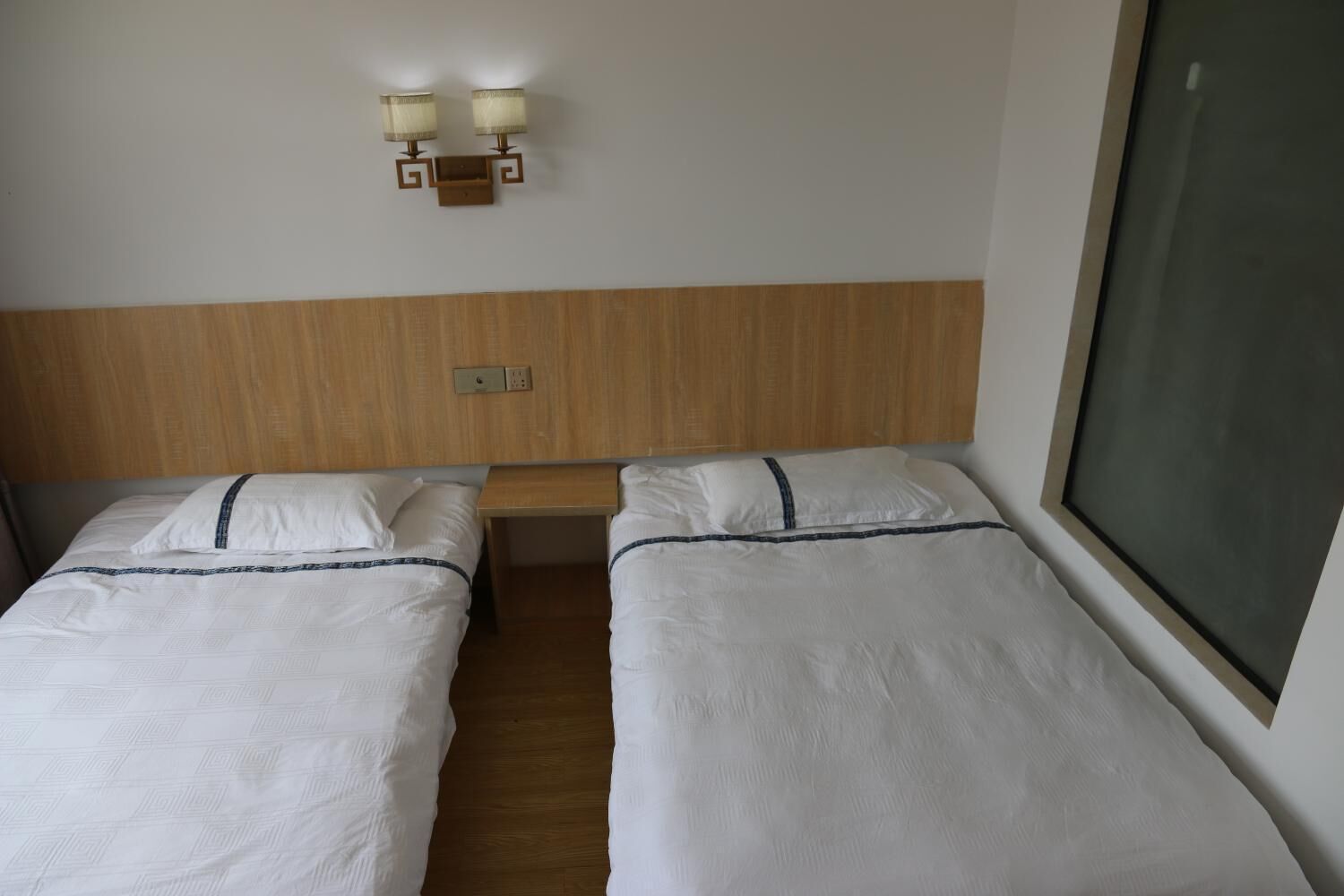A hotel room with two single beds, a wooden headboard, a nightstand, and a wall-mounted lamp.