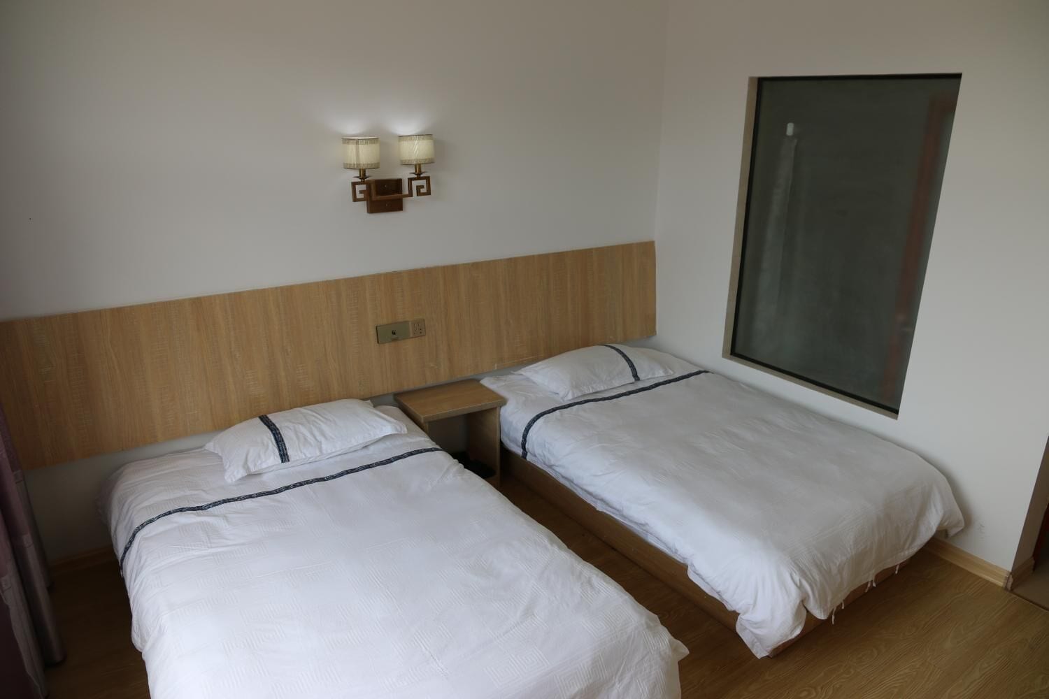 A hotel room with two single beds, a wooden headboard, a nightstand, and a wall-mounted light fixture.