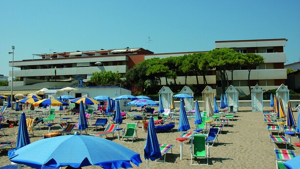 A beach with rows of beach chairs and umbrellas, a building in the background, and a clear sky. - Casamare (Lignano Sabbiadoro)