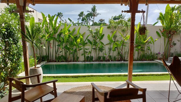 A poolside area with wooden furniture, a tiled floor, and lush greenery. - Villa Manik (Canggu)