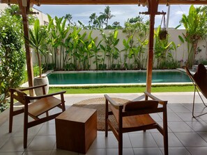A poolside area with wooden furniture, a tiled floor, and lush greenery. - Villa Manik (Canggu)