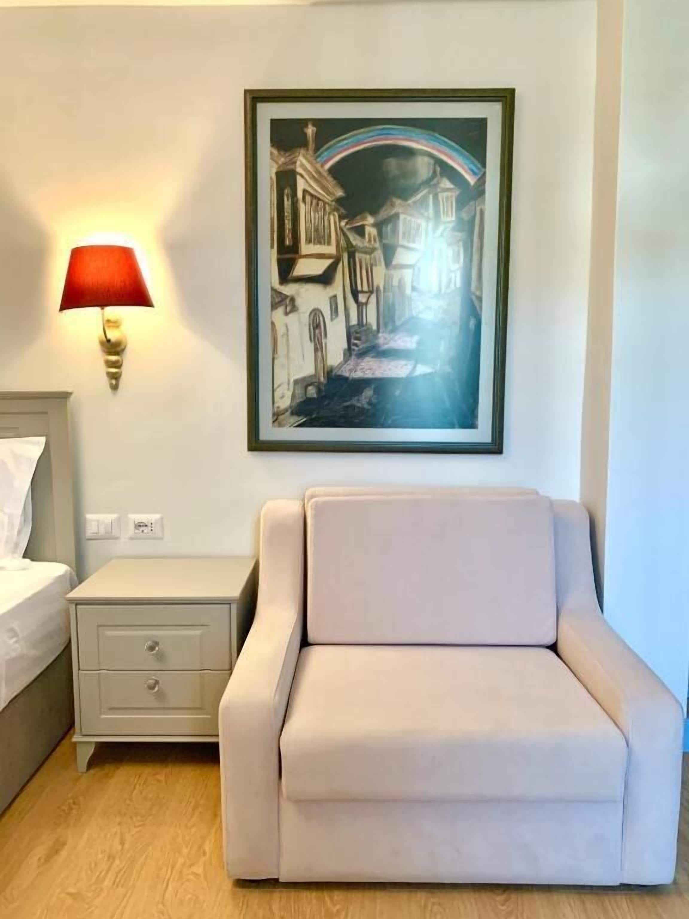 A hotel room with a white sofa, a nightstand with a lamp, and a framed picture of a street scene on the wall.