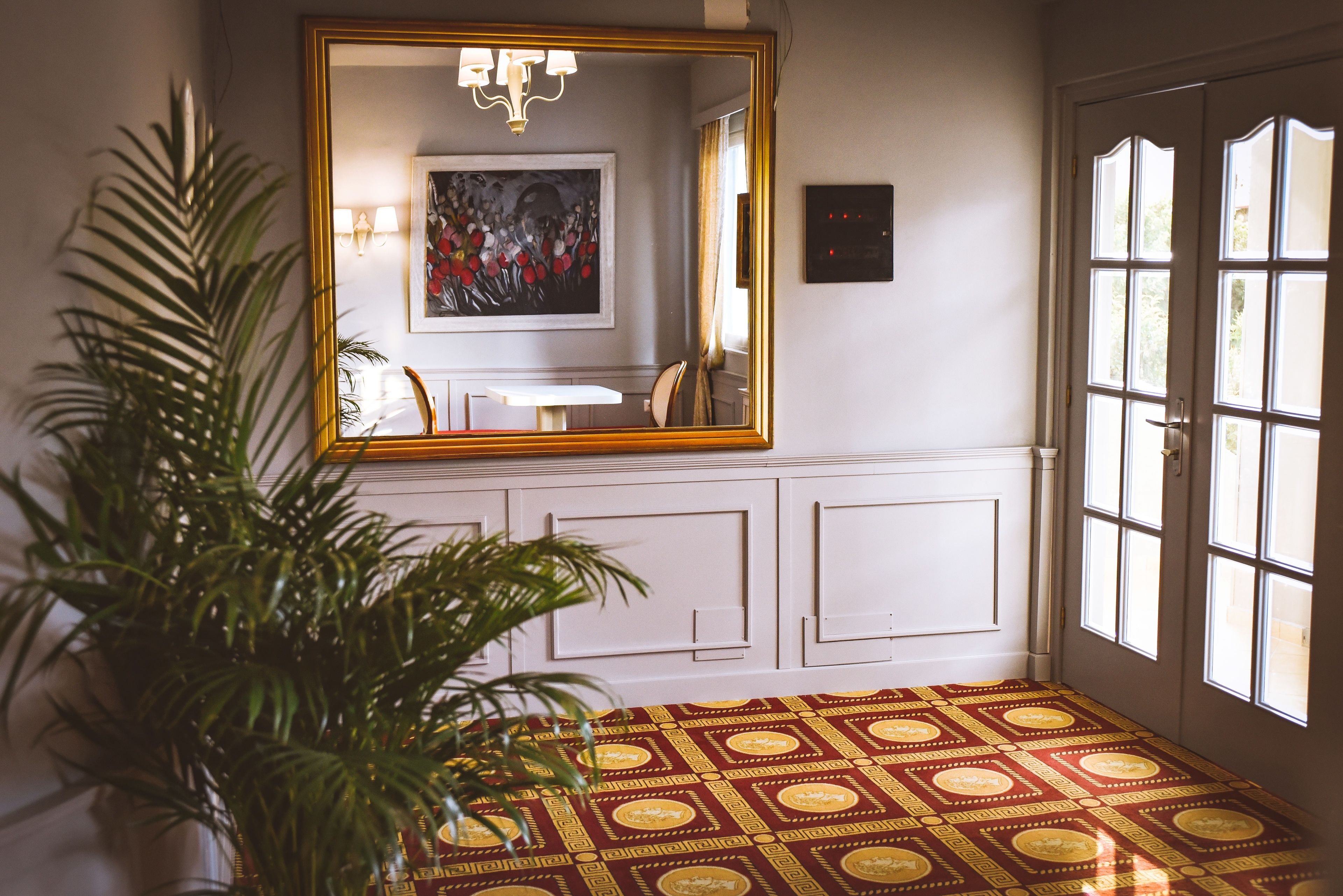 A room with a large mirror, a chandelier, a painting, a potted plant, and a patterned carpet.
