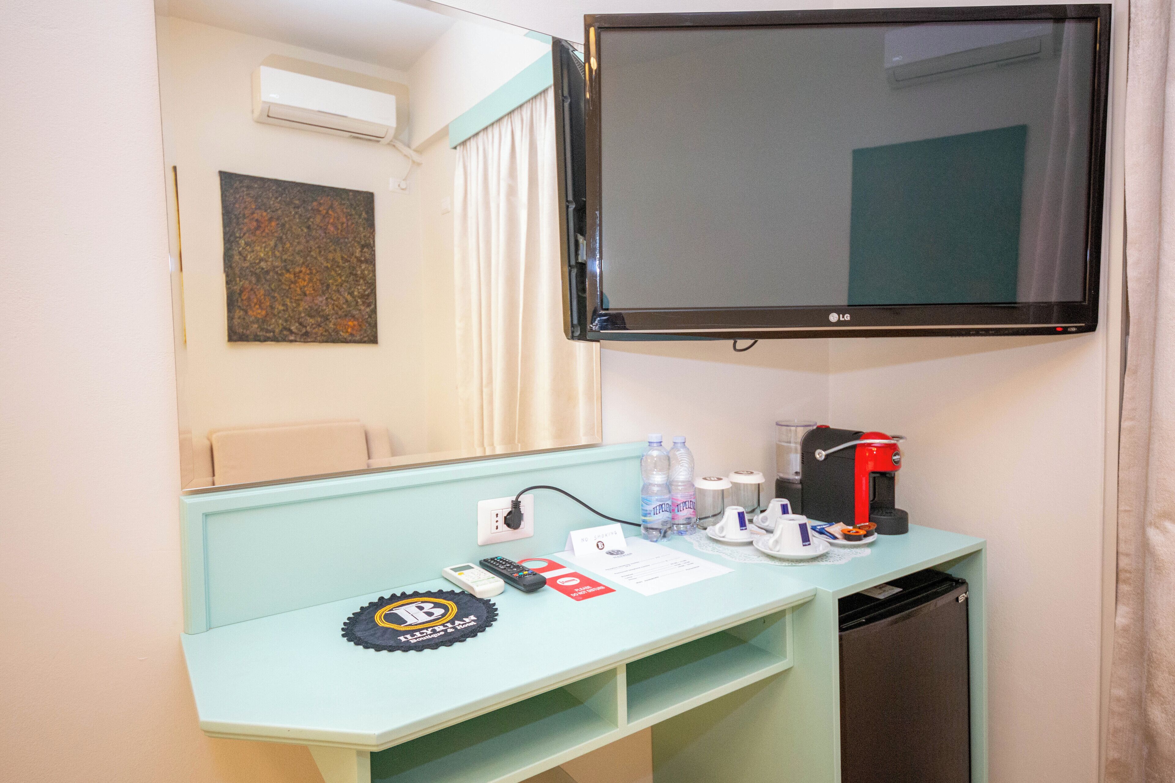 Habitación de hotel con una televisión montada en la pared, un pequeño escritorio con cafetera, botellas de agua y un control remoto.