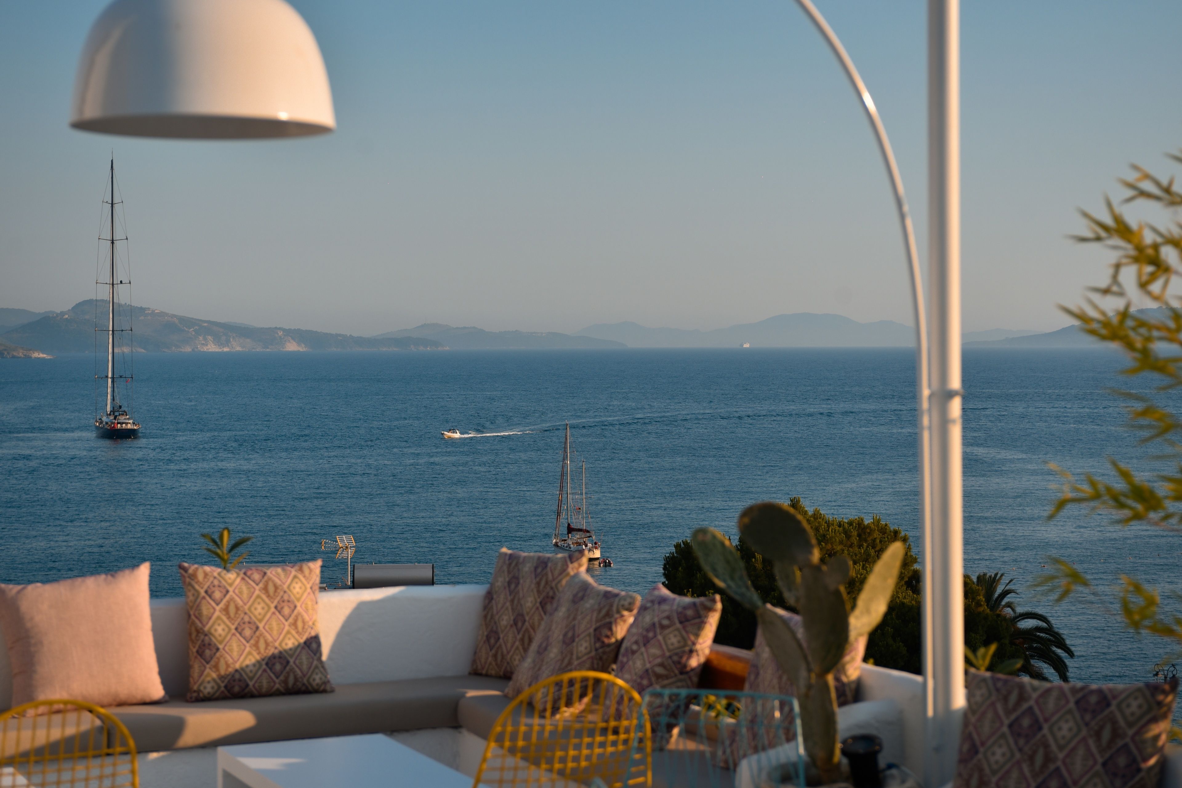 A seaside terrace with a sofa, cushions, and a lamp, overlooking the sea with boats and distant hills.