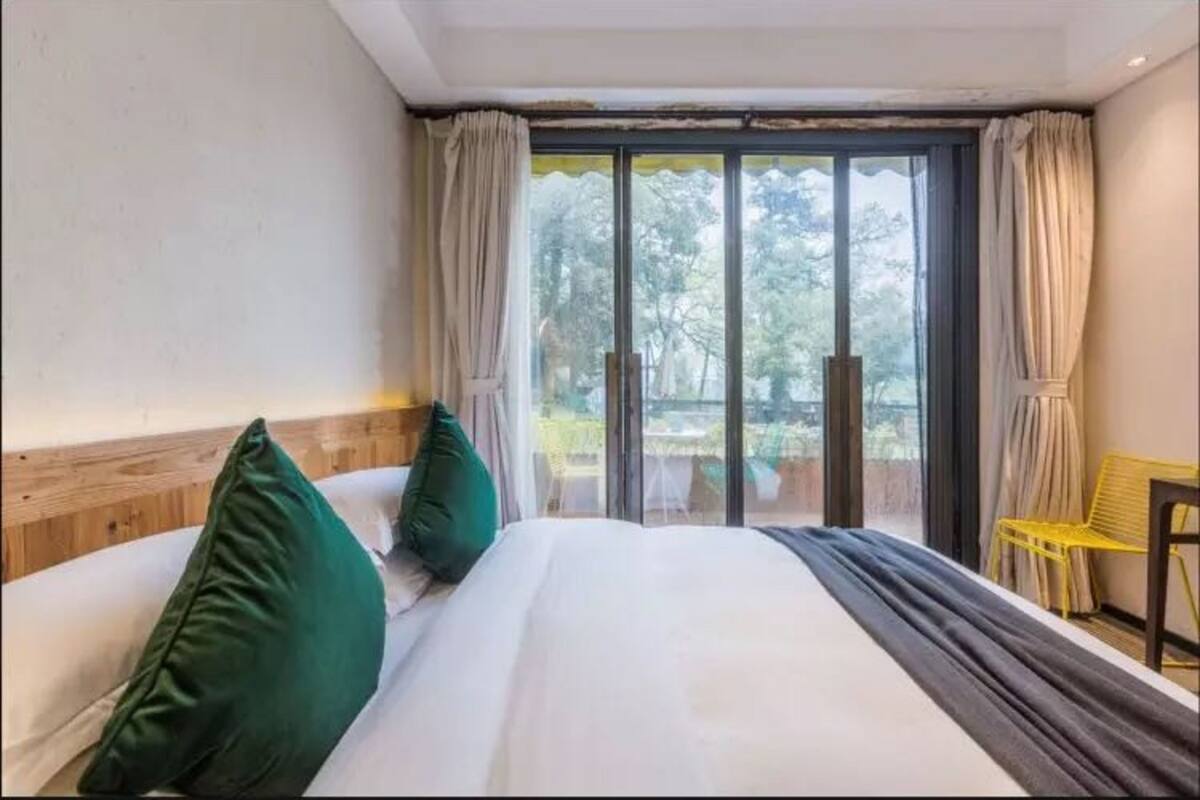 Ein Hotelzimmer mit einem großen Bett, zwei Kissen und Blick durch eine Glastür auf einen Außenbereich mit einem gelben Sessel.