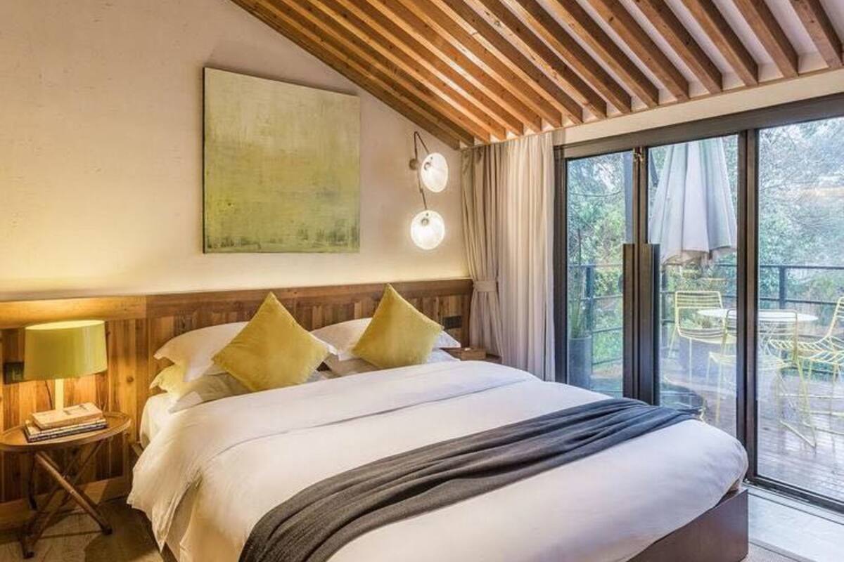 Ein Hotelzimmer mit einem großen Bett, einem Holzkopfstück, einem Nachttisch mit Lampe, einem Gemälde an der Wand und einem Balkon mit Sitzmöbeln.