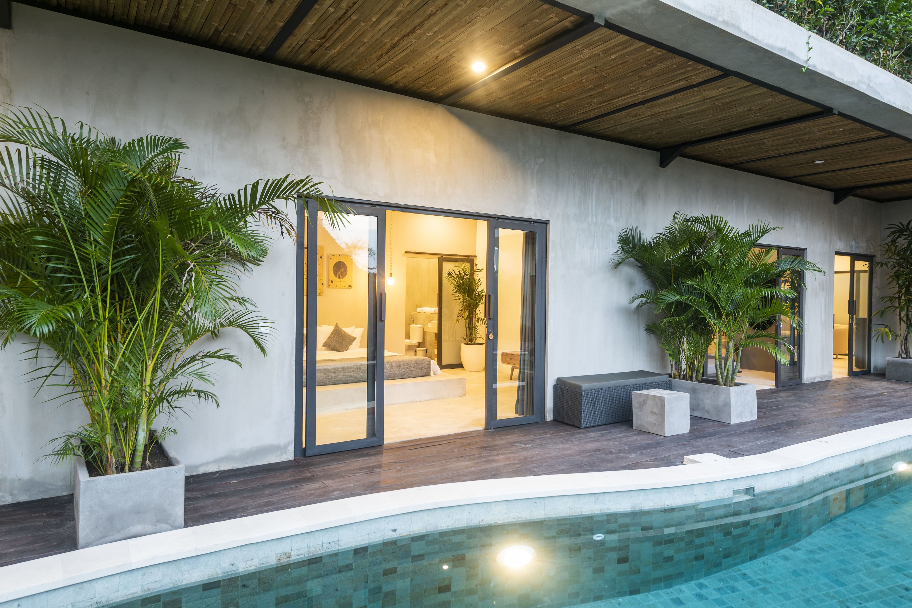 A modern poolside area with a built-in bench, potted plants, and a glass door leading to a bedroom.