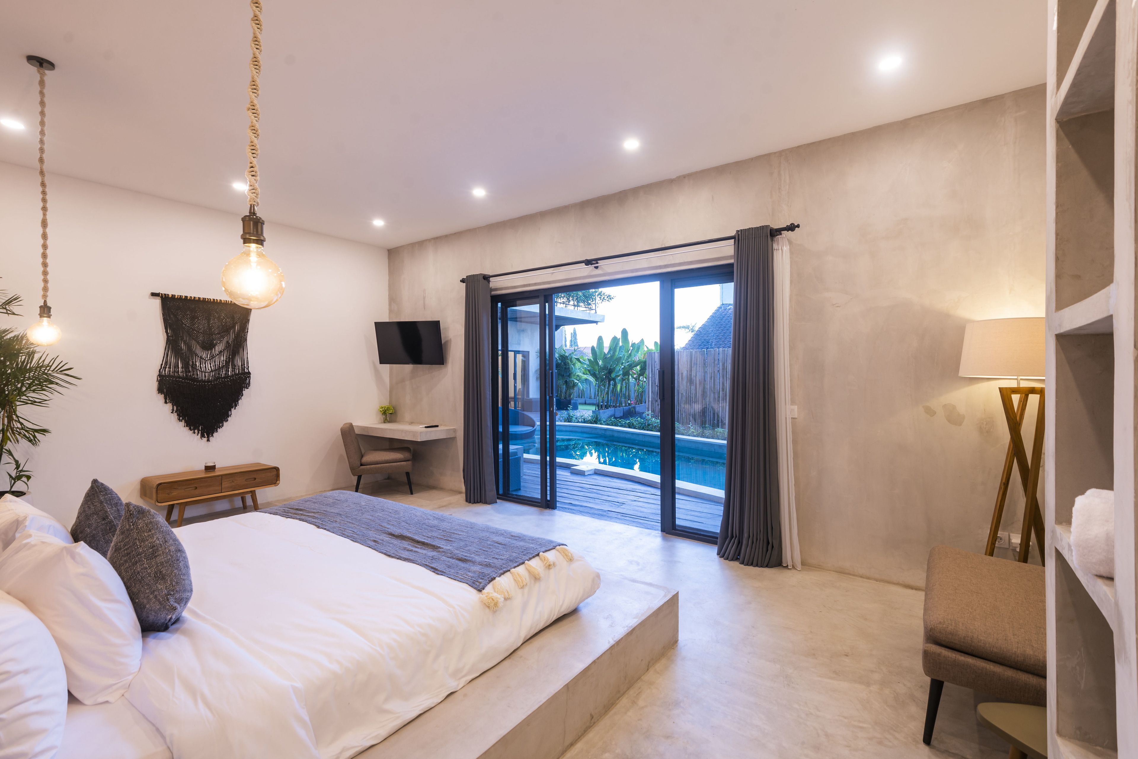 A modern bedroom with a large bed, a desk, a TV, and a view of a pool.