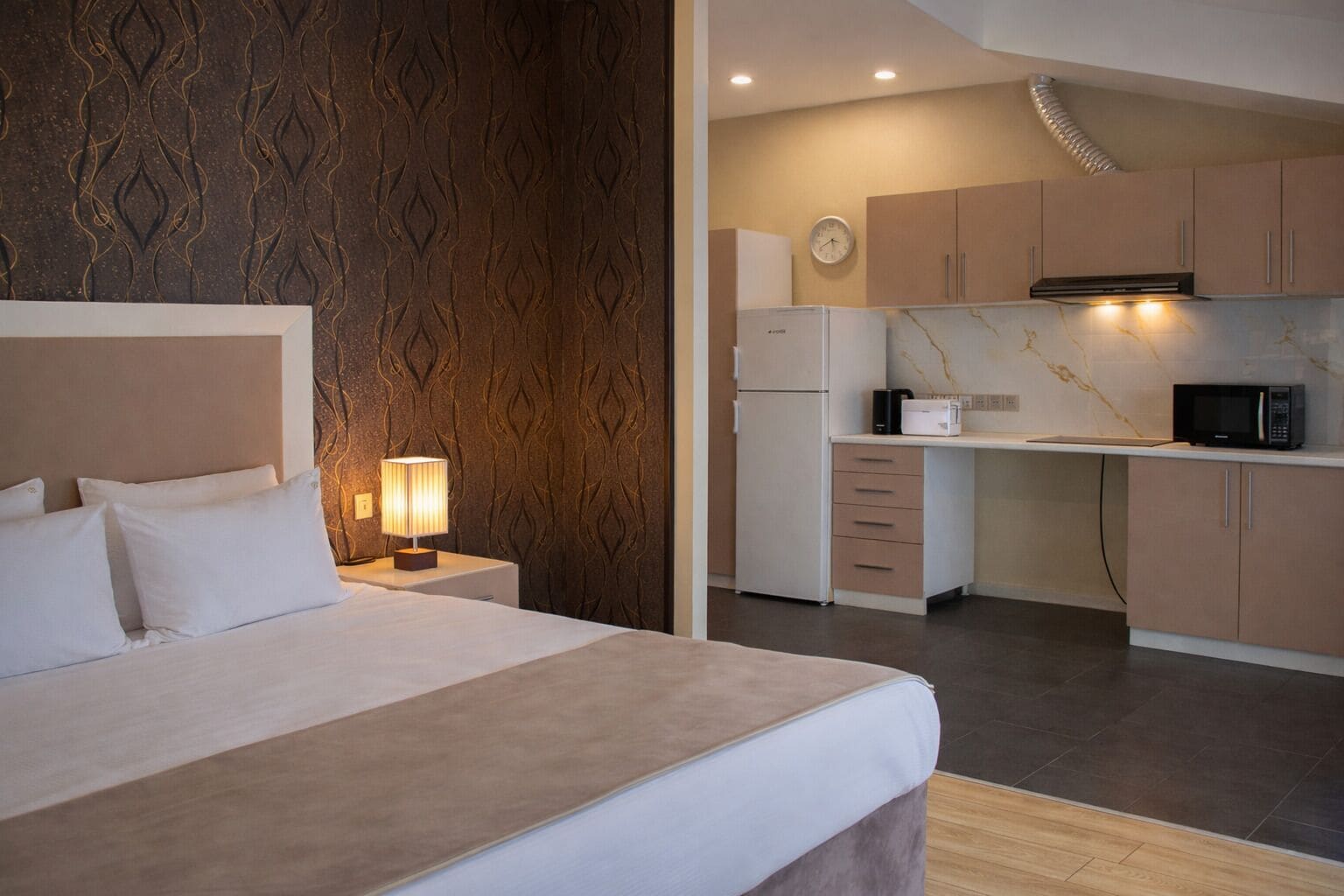 A hotel room with a bed, bedside lamp, and a kitchenette with a microwave, refrigerator, and cabinets.