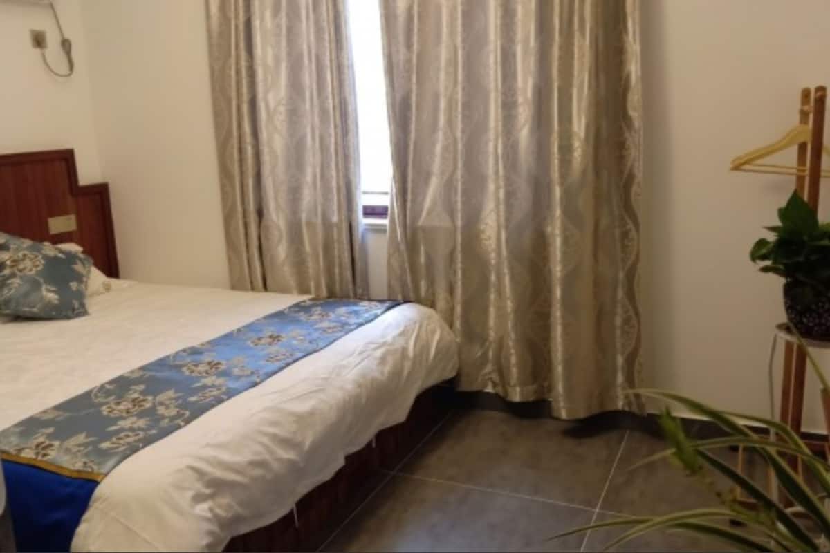 A hotel room with a bed, curtains, a plant, and a wooden headboard.