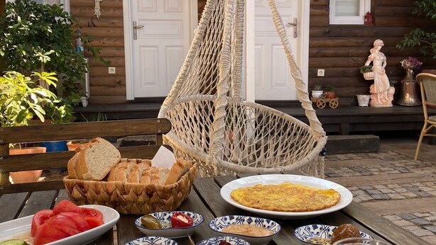 A wooden table with a variety of dishes, a hanging macrame chair, and a rustic building in the background. - Alacati Sardunya Hotel (Cesme)