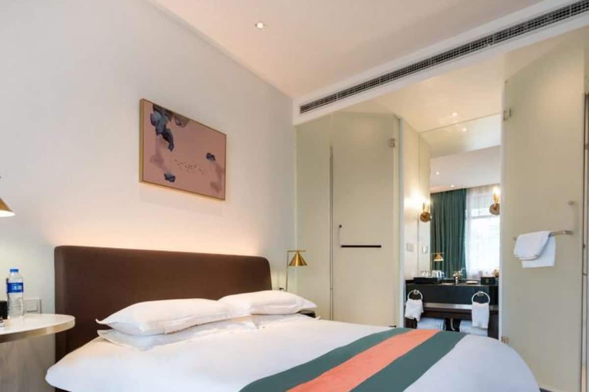 A hotel room with a large bed, a bedside table with a lamp, a painting on the wall, and a bathroom visible through an open door.