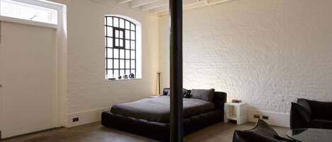 A bedroom with a large bed, a small bedside table, a window with bars, and a white brick wall.