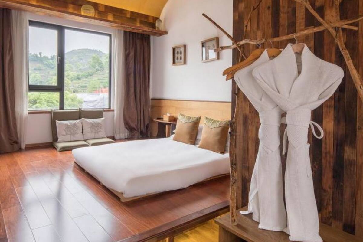 Un dormitorio con una cama grande, suelo de madera, vista a la vegetación exterior y una pared de madera con un adorno de rama.