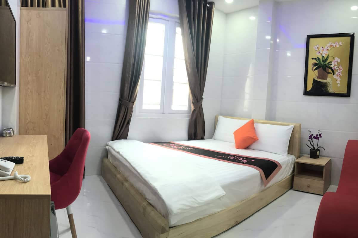 A hotel room with a bed, a desk with a telephone, a chair, a bedside table with a plant, and a window with curtains.