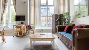 A modern living room with a sofa, a coffee table, a TV, and a view of a cityscape through large windows. - Bright Two BR Period Apartment in Whitechapel (London)