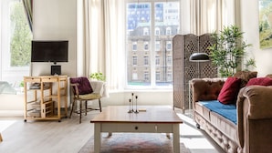 A modern living room with a sofa, a coffee table, a wooden side table, a television, and a view of buildings through large windows. - Bright Two BR Period Apartment in Whitechapel (London)