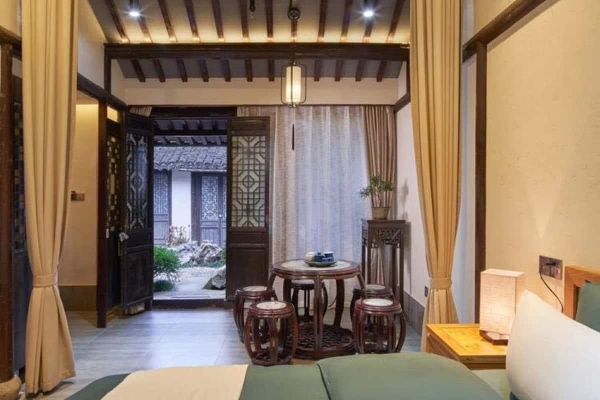 A traditional-style bedroom with a bed, a small round table with stools, a nightstand, and a view of a garden through an open door.