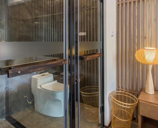 Une salle de bain moderne avec une porte en verre, des toilettes blanches, une étagère en bois et une chaise en osier.