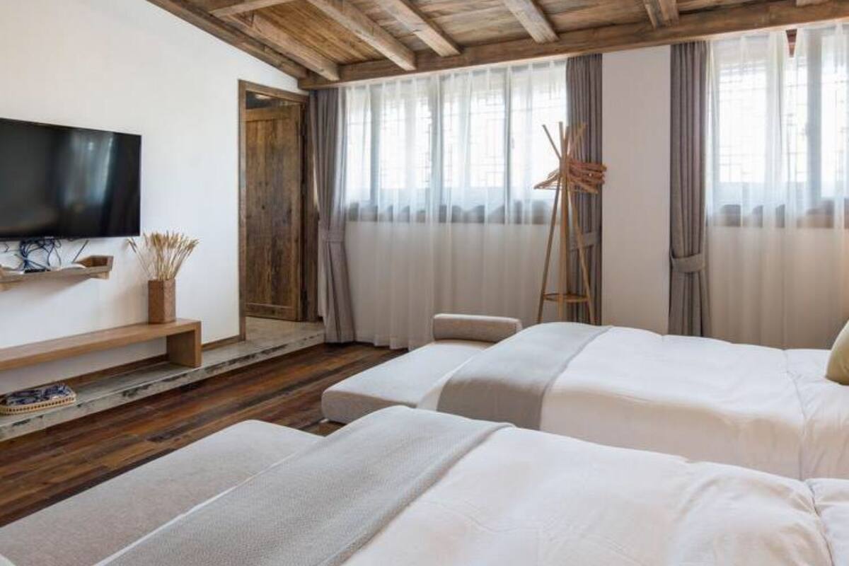 A hotel room with wooden beams on the ceiling, a flat-screen TV mounted on the wall, two beds with white bedding, a wooden side table, and a window with sheer curtains.