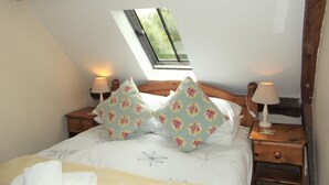 A bedroom with a sloped ceiling, a skylight, a bed with floral pillows, two bedside tables with lamps, and a wooden headboard. - Bridge Farm Holiday Cottages (Evesham)