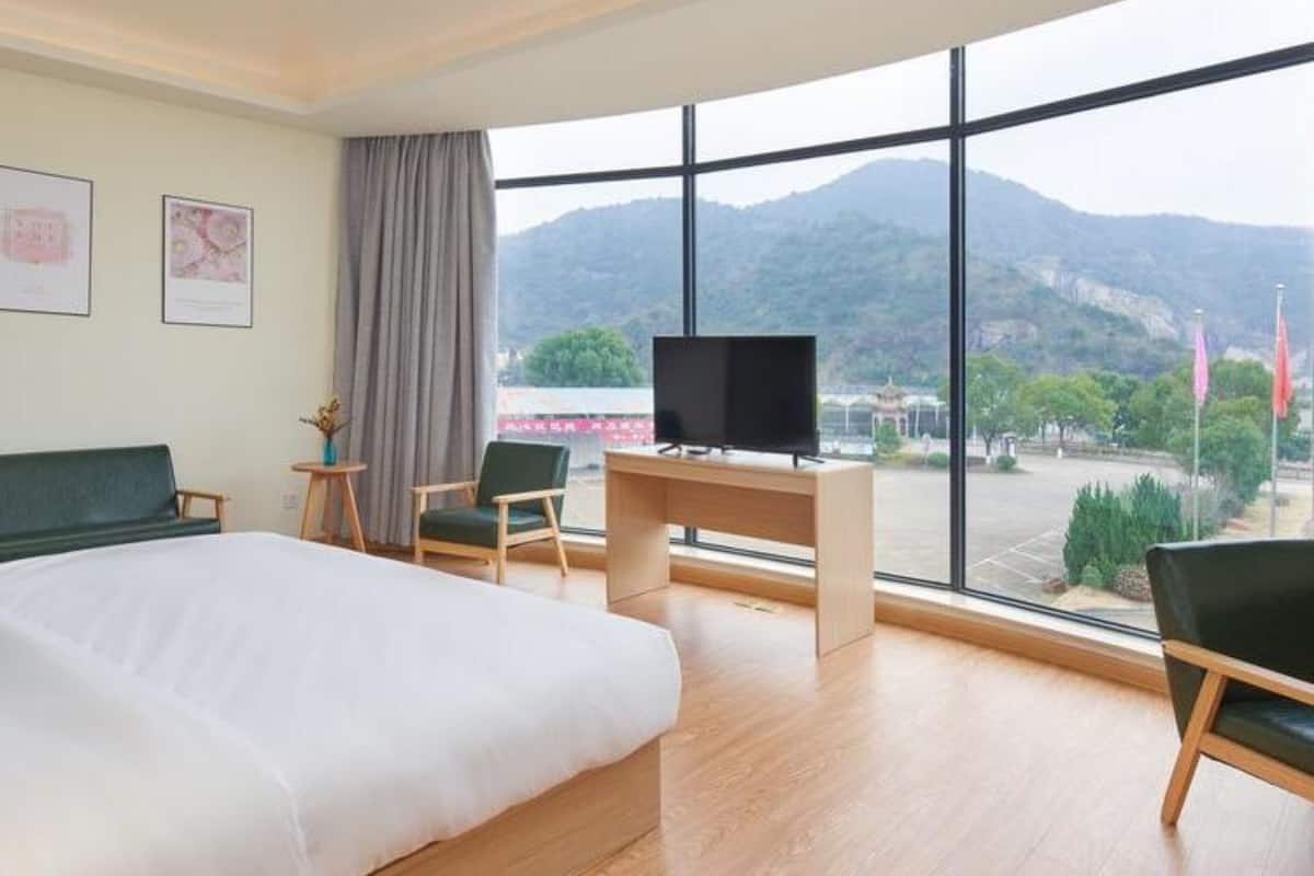 Quarto de hotel com uma cama grande, uma mesa de trabalho com televisão e vista para montanhas através de amplas janelas.