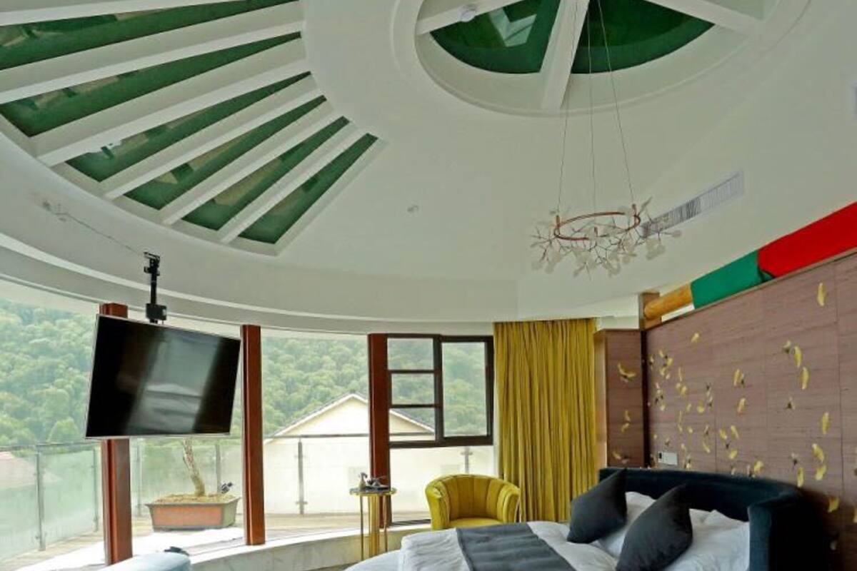 Un dormitorio con una cama curvada, un televisor montado en la pared, un banco y vistas a la vegetación exterior.