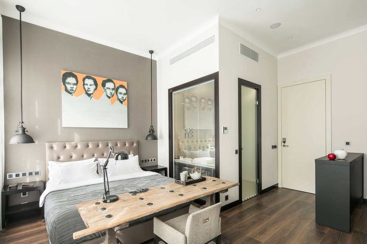 A modern hotel room with a large bed, a wooden desk, a chair, a wardrobe, and a mirror.